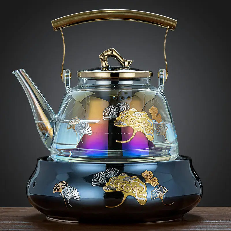 Glass tea kettle with gold ginkgo leaf pattern, gold handle, and matching black ceramic warmer stove base.