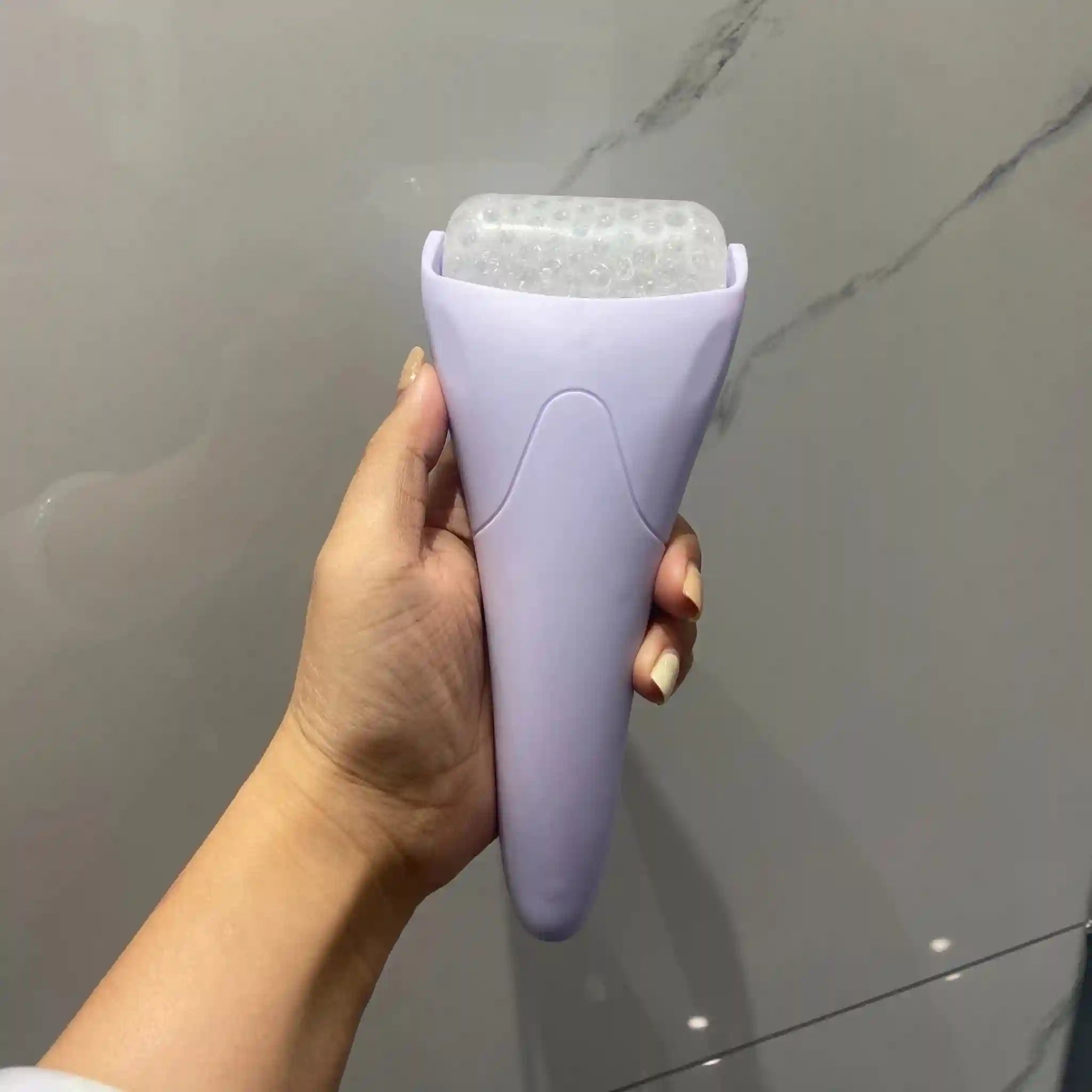 Handheld lilac facial ice roller with a textured, translucent cooling drum held in a person's hand.
