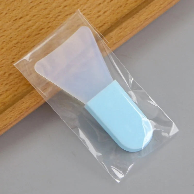 A clear, flexible silicone facial mask applicator with a light blue handle, packaged in a small plastic sleeve.