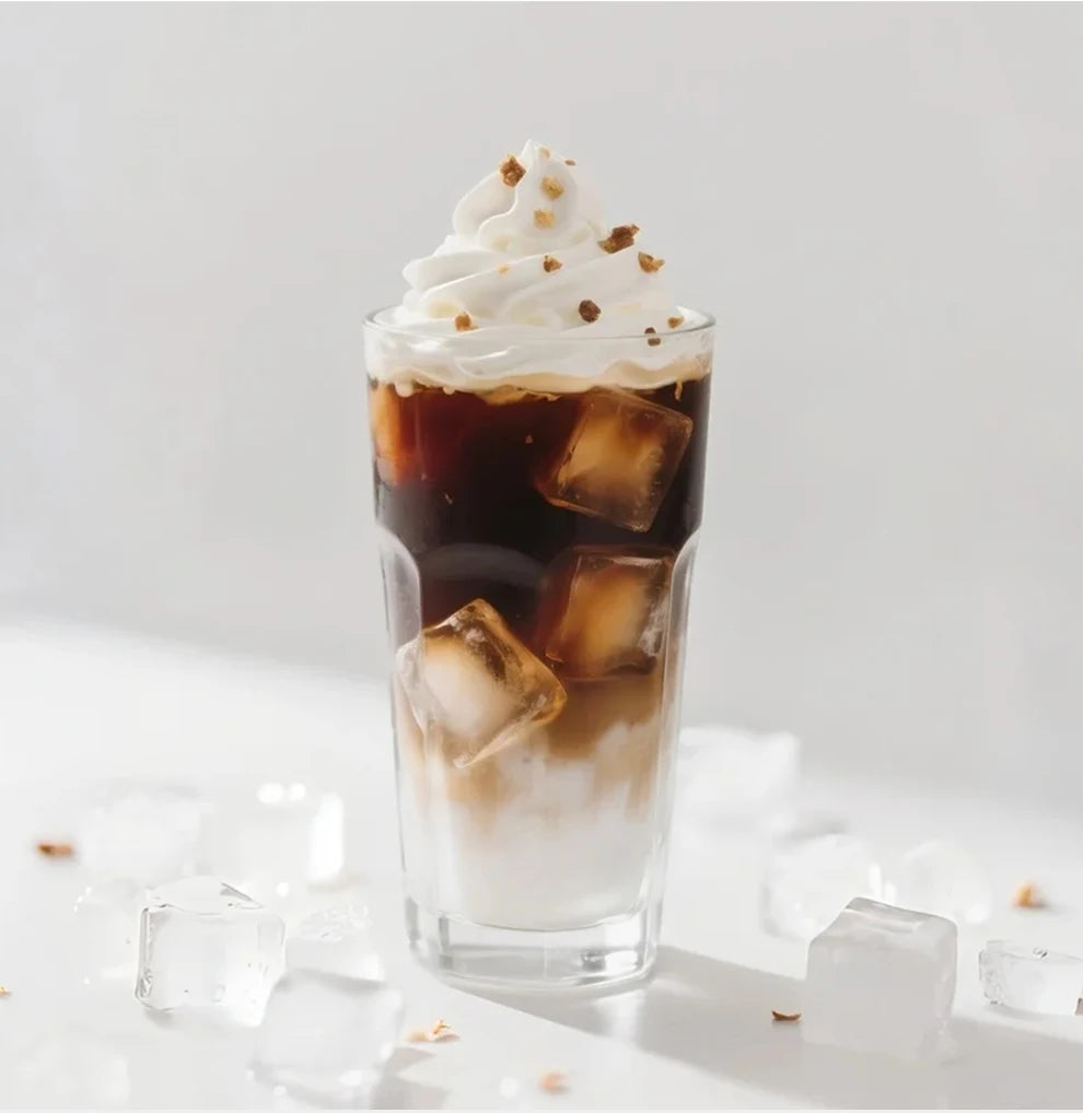 Glass of iced coffee with milk, whipped cream, and toppings, served in a clear tall drinking glass on a white surface.