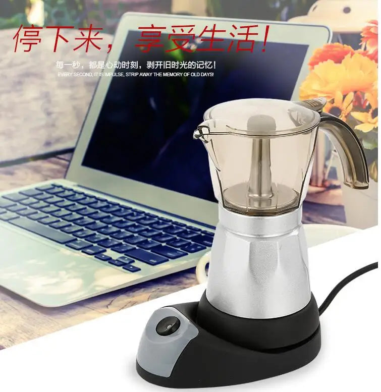 Electric moka pot coffee maker with transparent top, silver aluminum base, and black heating stand on office desk.