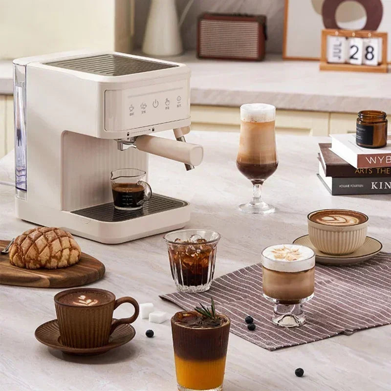 Compact white espresso machine with portafilter on a marble kitchen counter next to various coffee drinks and pastry.