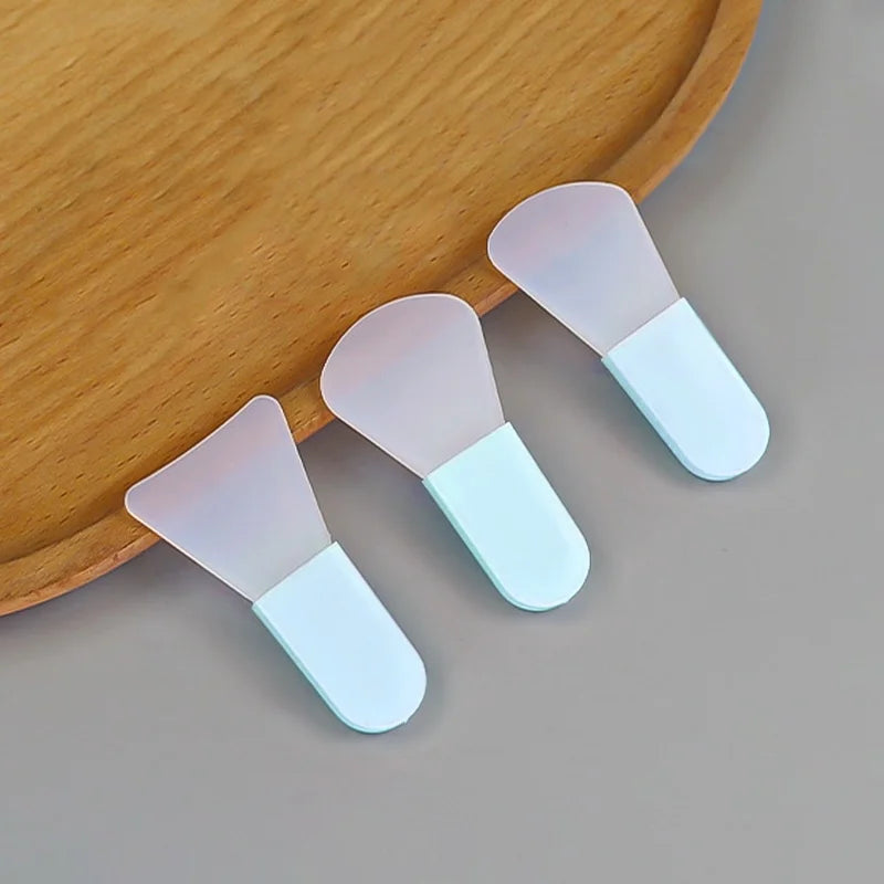 Three silicone facial mask brushes with translucent flexible heads and solid light-blue handles on a wooden tray.