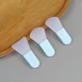 Three silicone facial mask brushes with translucent flexible heads and solid light-blue handles on a wooden tray.