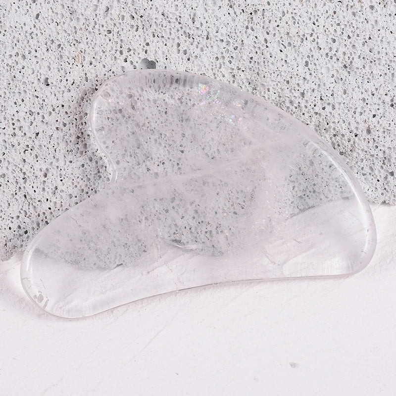 A heart-shaped, clear translucent crystal gua sha facial massage tool resting on a textured gray stone surface.