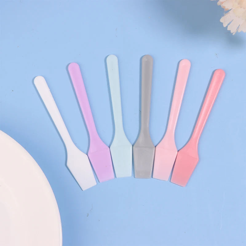 Six colorful mini plastic cosmetic spatulas arranged in a row against a light blue background.