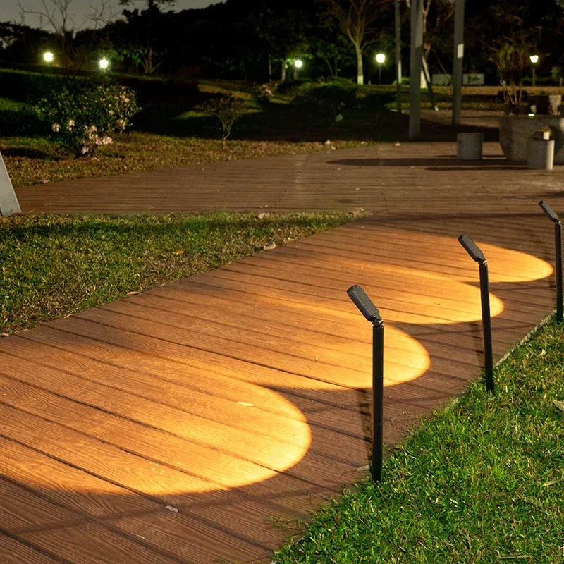 Four black LED solar spotlights line a wooden walkway, casting warm pools of light on the planks.