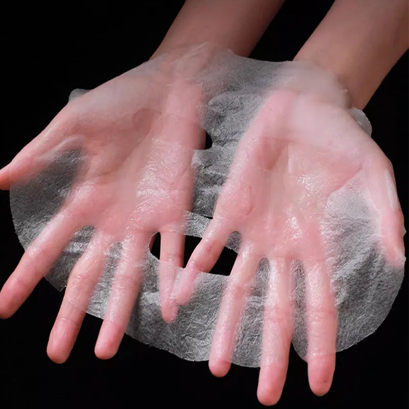 Two hands hold a thin, translucent, white disposable facial mask sheet against a black background.