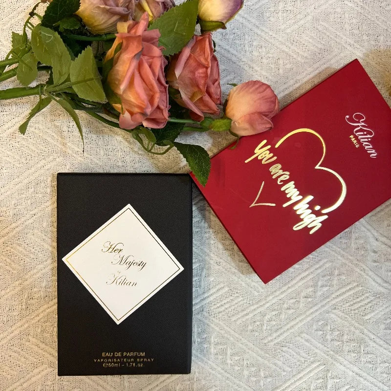 Her Majesty 50ml eau de parfum in a rectangular black box with a gold-trimmed label next to a red gift box and roses.
