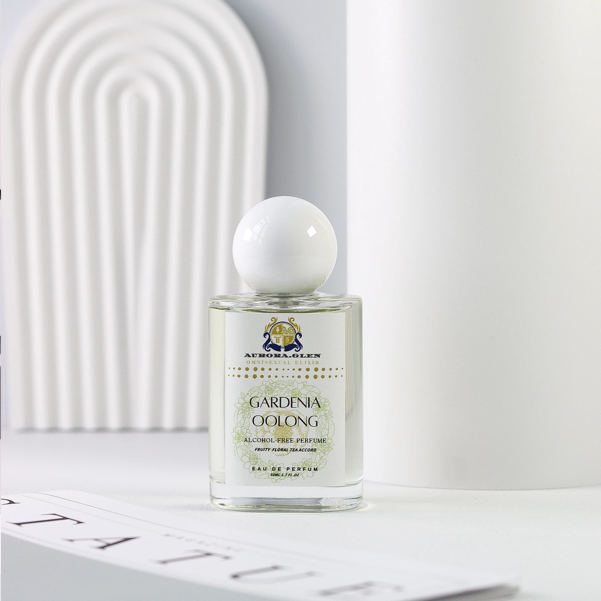 Glass perfume bottle with white spherical cap, Gardenia Oolong label, clear liquid fragrance on white minimalist surface.