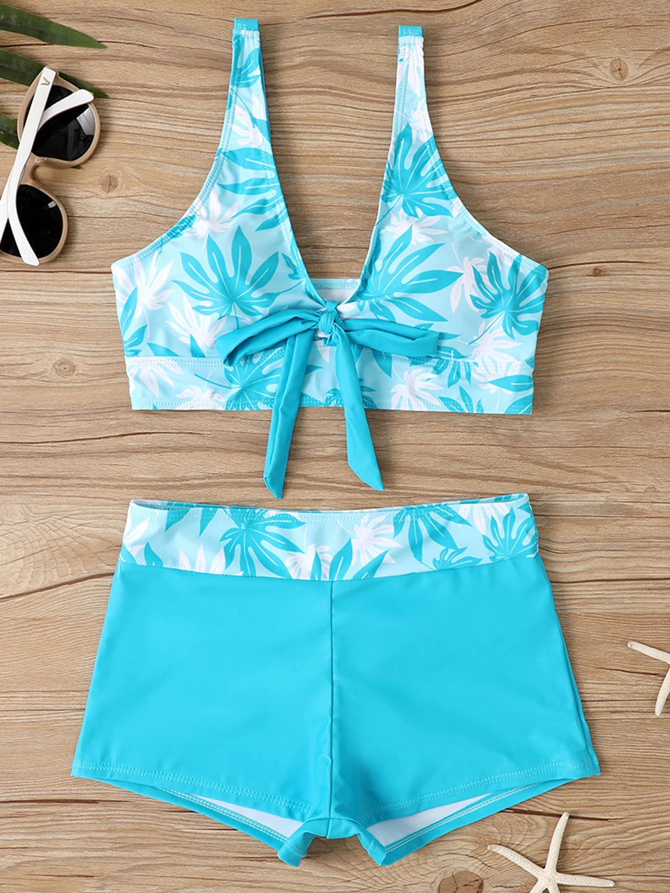 Tidesmit turquoise leaf-print high-waisted bikini with a front-tie bralette top and solid bottoms on a wood background.