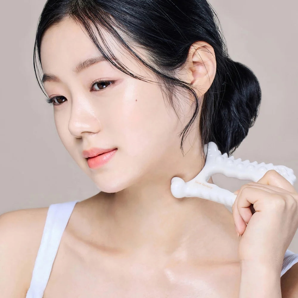 A woman using a white, textured ceramic Y-shaped gua sha tool to massage her neck and jawline.
