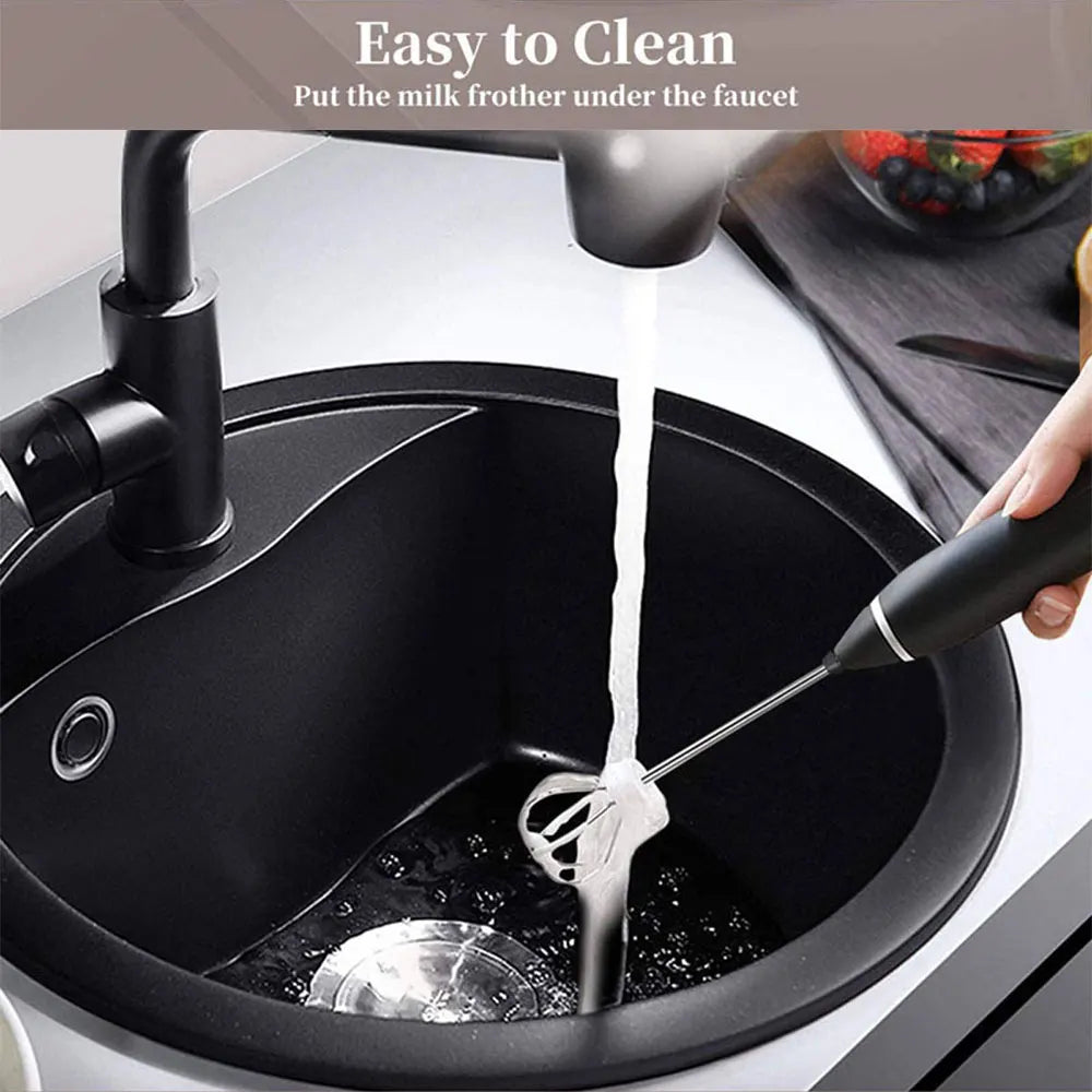 Handheld black electric milk frother with stainless steel whisk being rinsed under a kitchen faucet for easy cleaning.