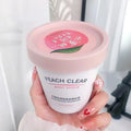 Peach Clear body scrub in a small, white cylindrical container with a pink lid featuring a peach graphic.