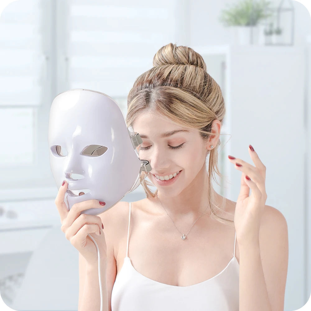 A woman holds a smooth, white, contoured LED light therapy face mask in a bright, modern room.