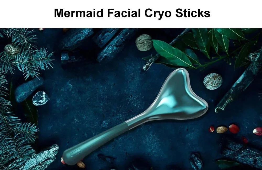 A mermaid-tail shaped metallic facial cryo stick rests on a dark surface surrounded by pine branches and nuts.