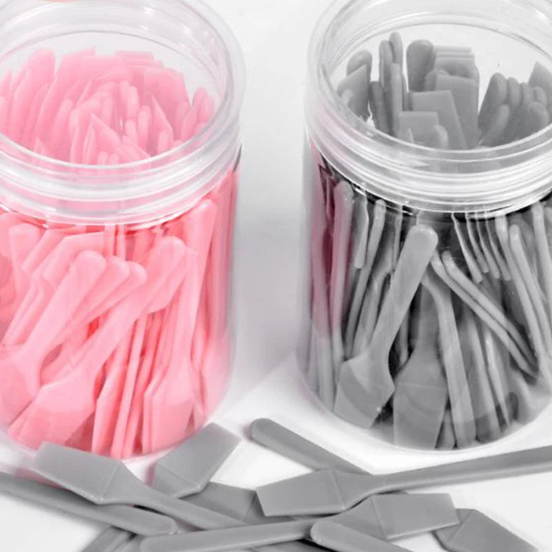 Two clear jars filled with many small, flat pink and grey plastic cosmetic spatulas for applying creams and masks.