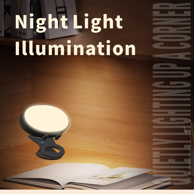 Small, round, black clip-on LED reading light casting a warm glow onto an open book on a wooden desk.
