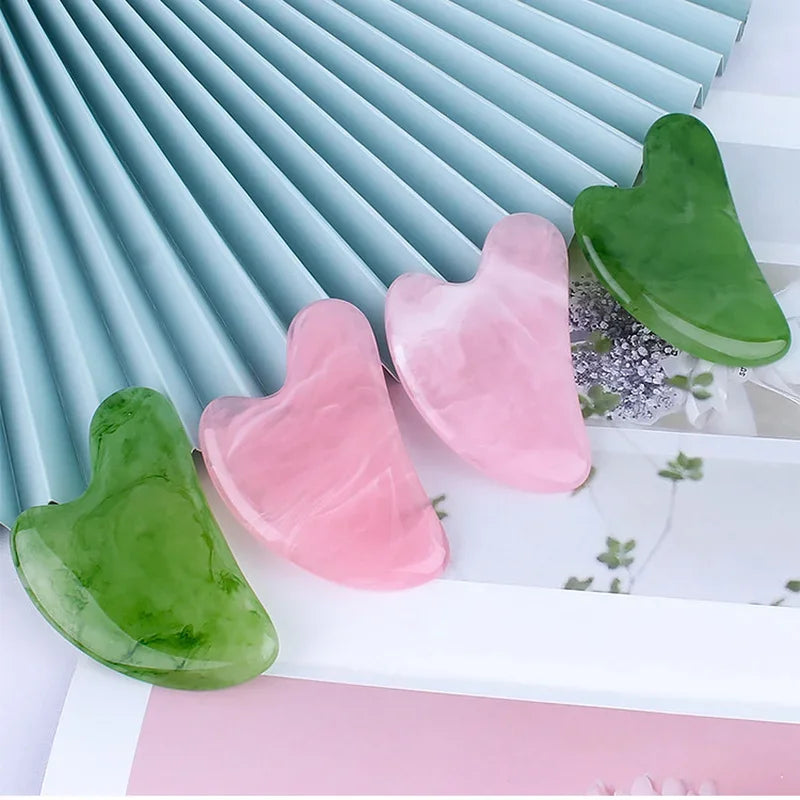 Four heart-shaped resin gua sha massage tools in shades of marbleized green and pink resting on decorative paper.