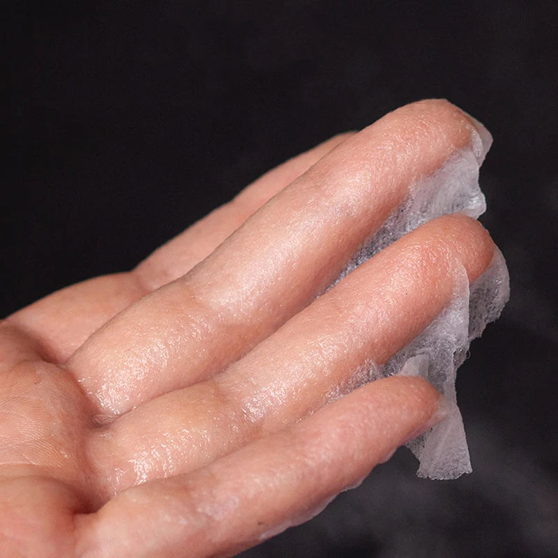 A person's hand holding a thin, translucent, white disposable facial mask sheet against a dark background.