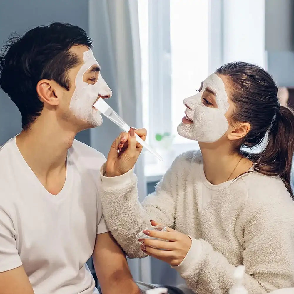 Woman applying a white facial mask to a man’s face using a transparent, flat-tipped cosmetic applicator brush.