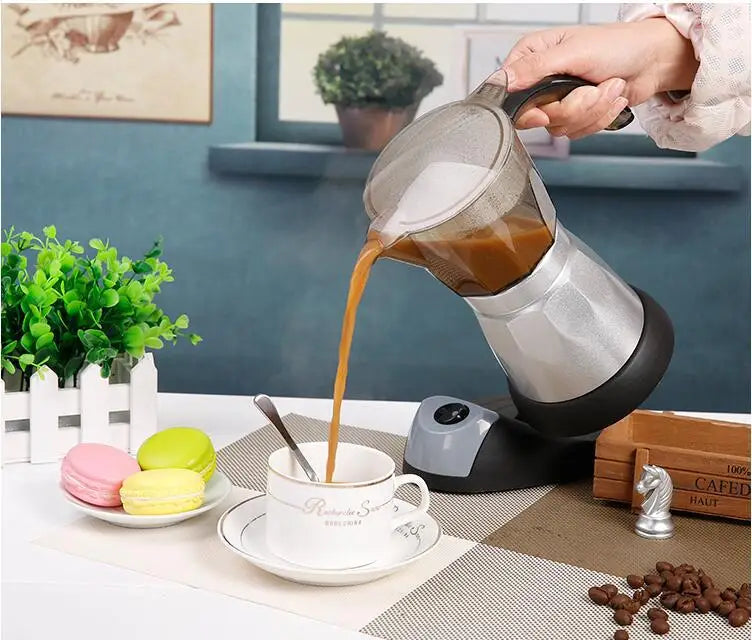 Electric silver aluminum moka pot pouring coffee into a white ceramic cup on a kitchen table.