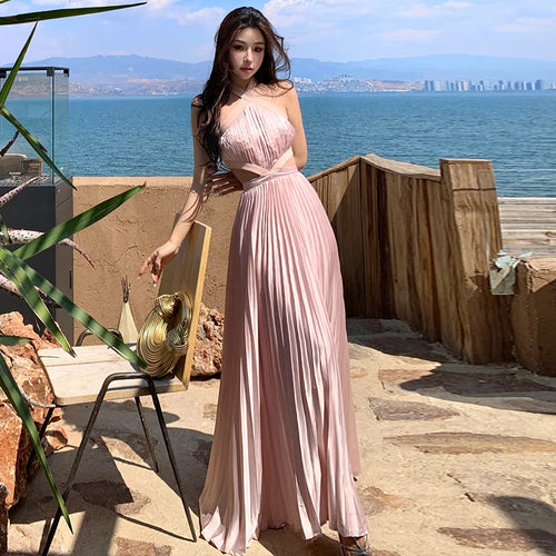 Tidesmit light pink satin halter neck maxi dress with a pleated skirt and cut-out waist, posing by the sea.