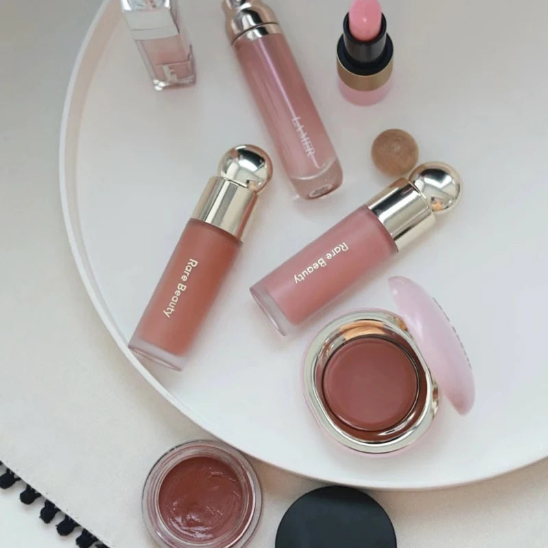 Rare Beauty makeup collection featuring silky liquid blush, luminizer, and cream lip products in neutral pink and nude.