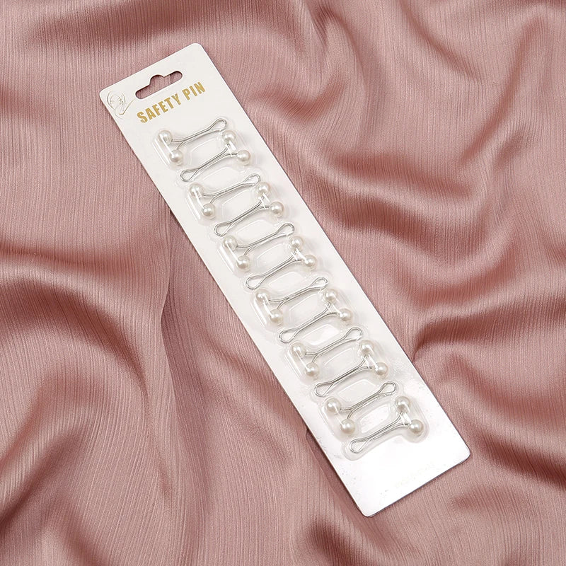 Pack of 12 silver metal hijab safety pins with white faux pearl ends, displayed on a card against pink fabric.