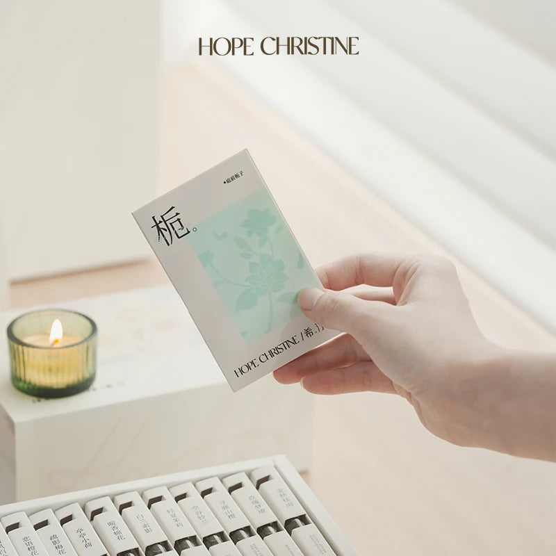 Hand holding Hope Christine perfume set, minimalist white box with floral packaging and a lit green candle nearby.