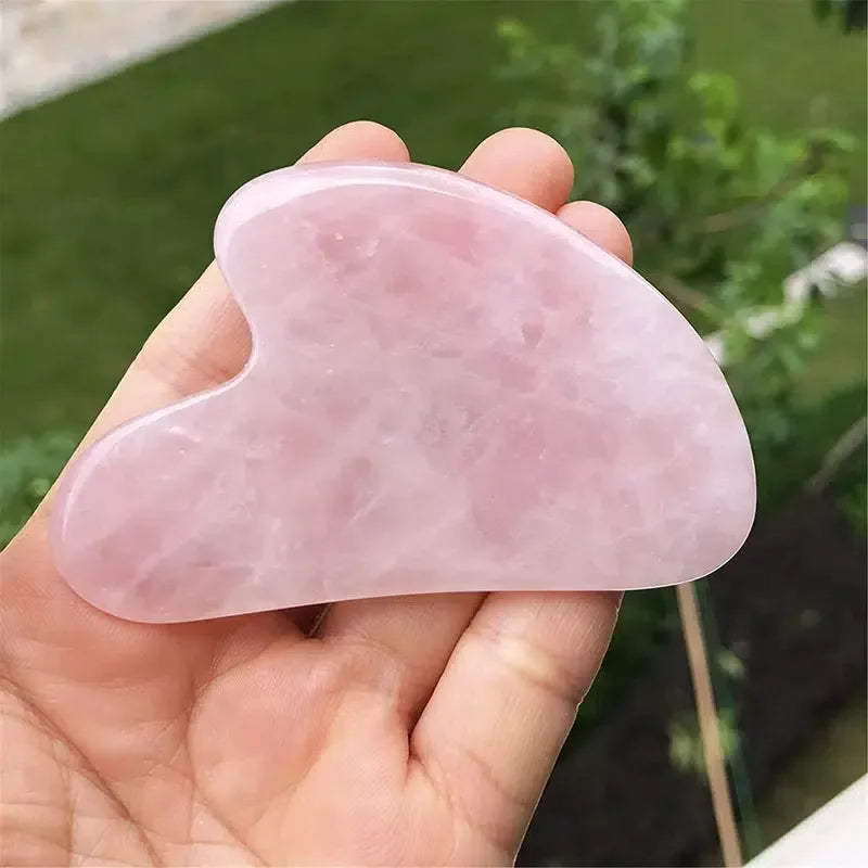 Hand holding a smooth, heart-shaped pink resin gua sha massage tool against a blurred outdoor background.