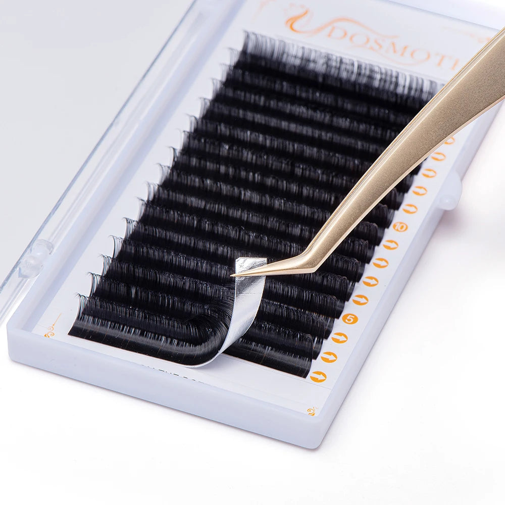 Black synthetic eyelash extension strips in a white tray, being lifted with gold tweezers for professional application.