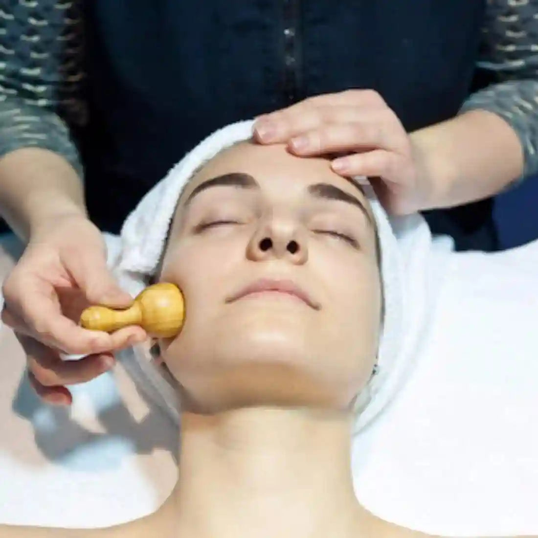A person receives a facial massage with a small, smooth, light-brown wooden mushroom-shaped tool.