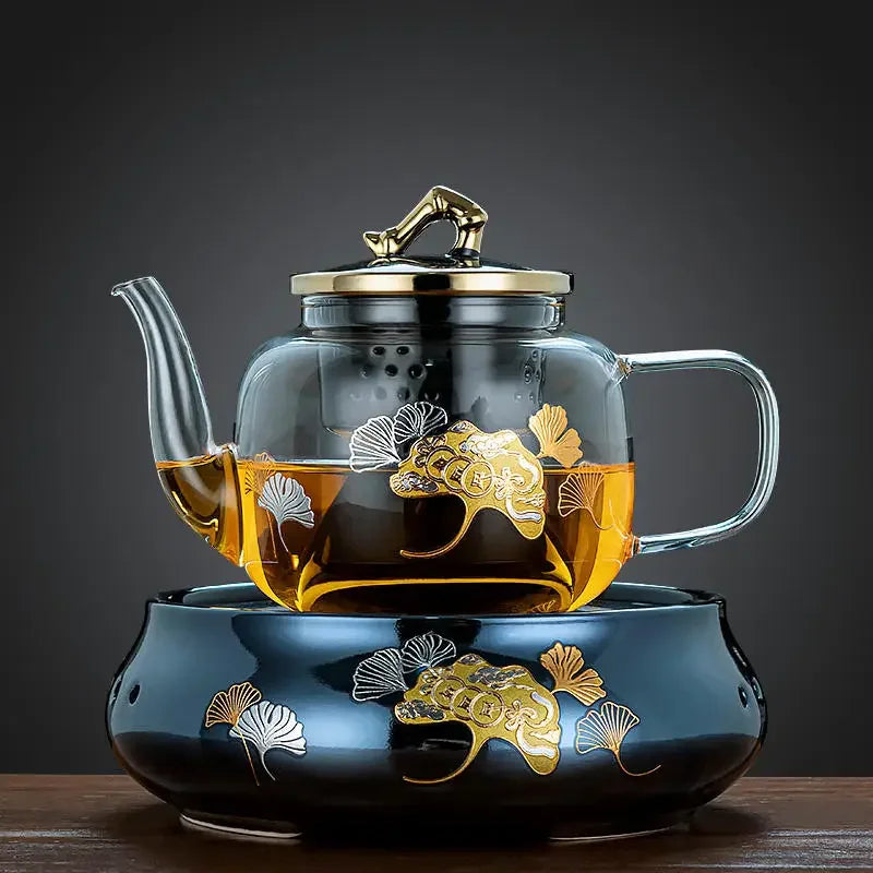 Clear glass teapot with gold ginkgo leaf design, metal lid, and matching ceramic warmer base on wooden table.