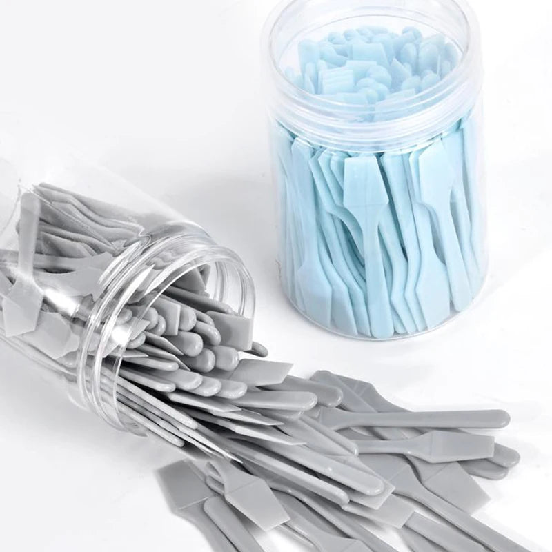 Two clear jars filled with many small, flat light blue and grey plastic cosmetic spatulas for skincare application.