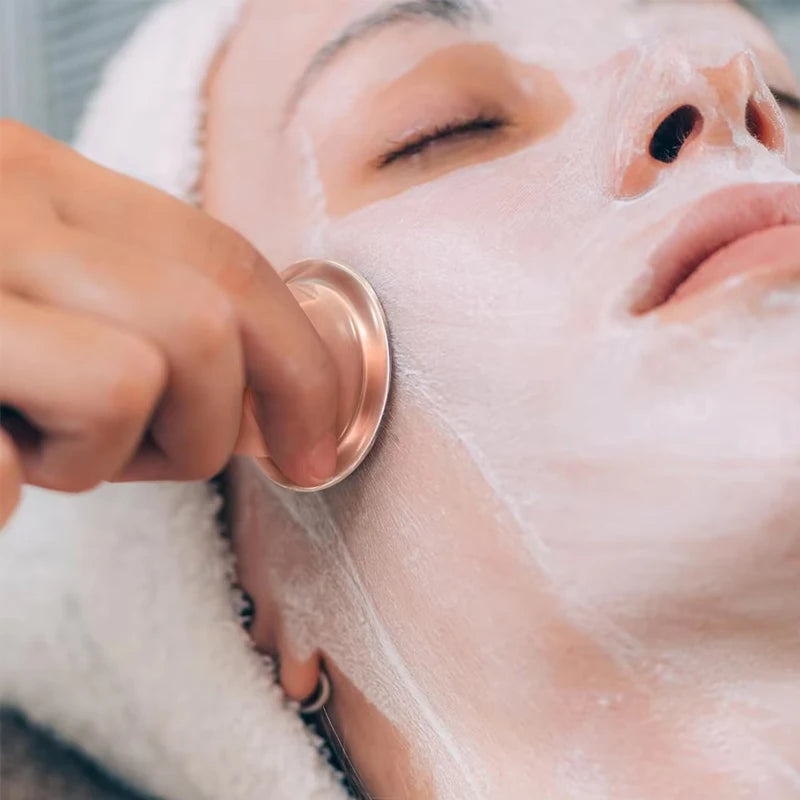 A hand using a small, metallic rose-gold facial cupping tool on a person's face covered in a white cream mask.