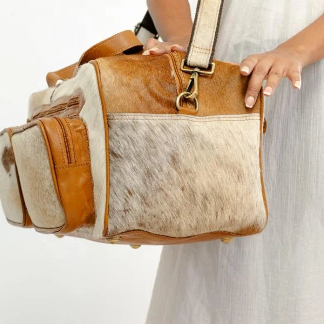 Western-style tan genuine leather and hair-on hide duffel trunk bag with side pockets and shoulder strap.