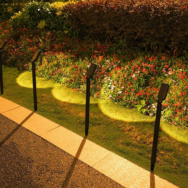 Four black LED solar spotlights illuminate a lawn beside a stone walkway.