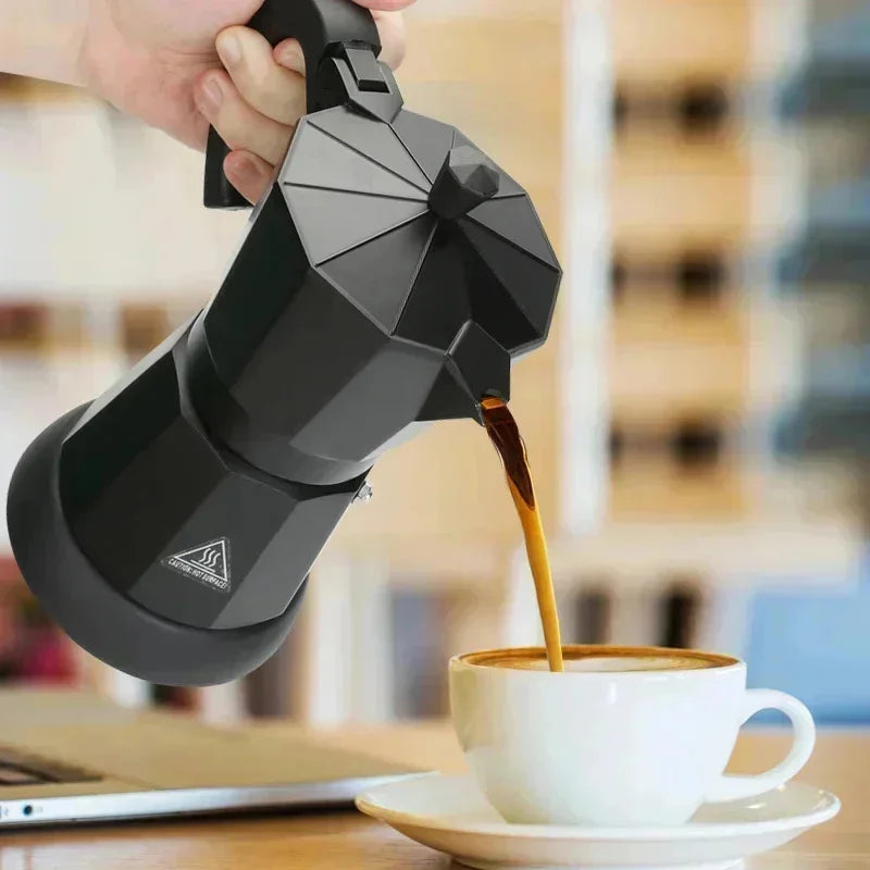 Hand pouring coffee from a modern matte black aluminum moka pot into a white cup on a wooden desk.
