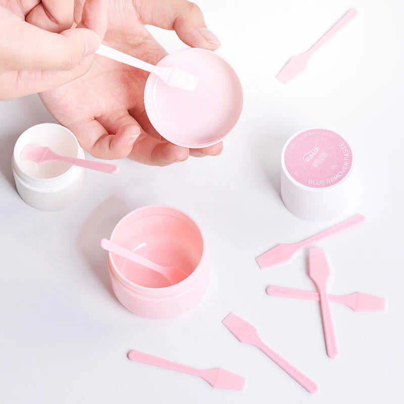 Set of small, light pink plastic cosmetic spatulas scattered around pink and white cream jars on a white background.