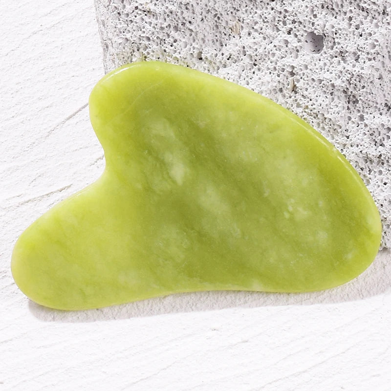 Smooth, heart-shaped light green jade gua sha facial massage tool resting on a textured white surface.