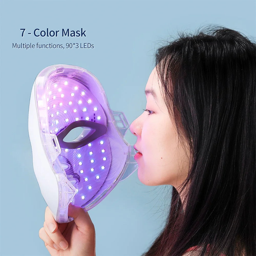A woman holds a transparent 7-color LED facial mask with visible lights against a light blue background.