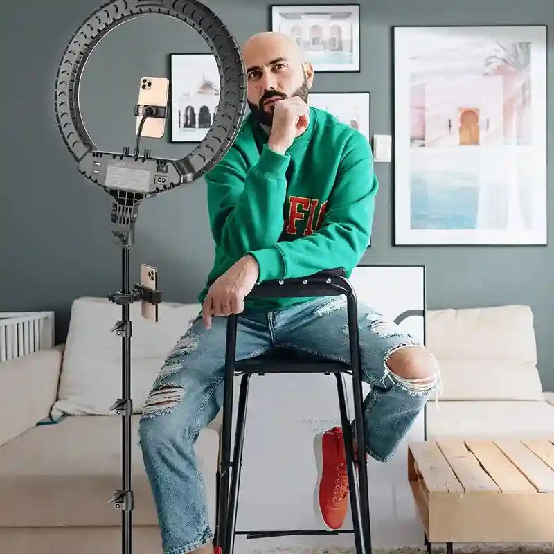 Black ring light on a tall tripod stand with phone mounts, used for recording video content in a home studio.