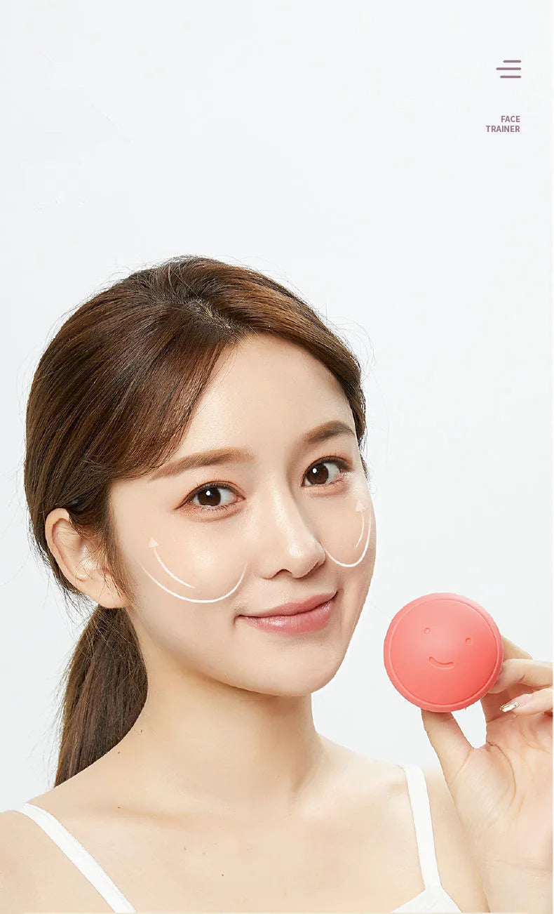 A woman holds a small, round, coral-pink silicone face-lifting exercise tool featuring a simple smiley face imprint.