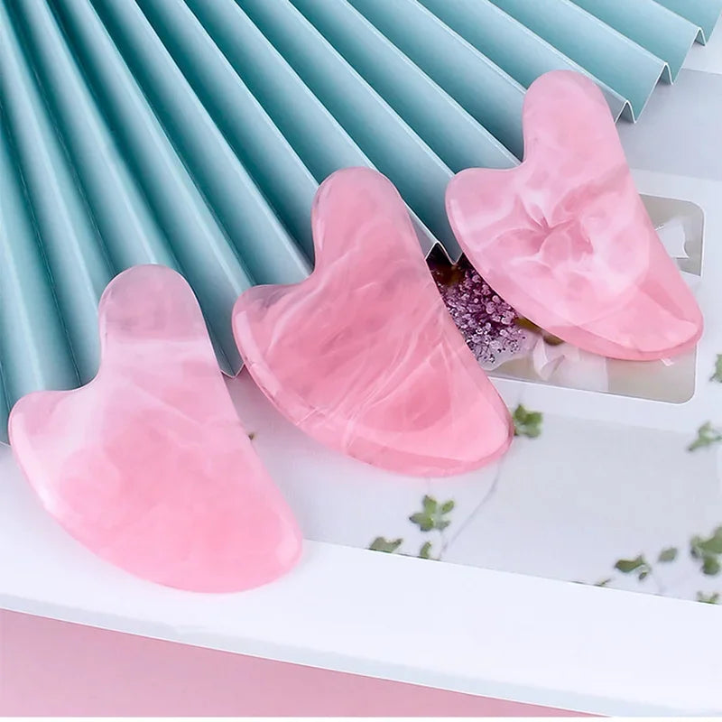 Three heart-shaped, pink marbled resin gua sha massage tools resting on a white surface with a blue pleated background.
