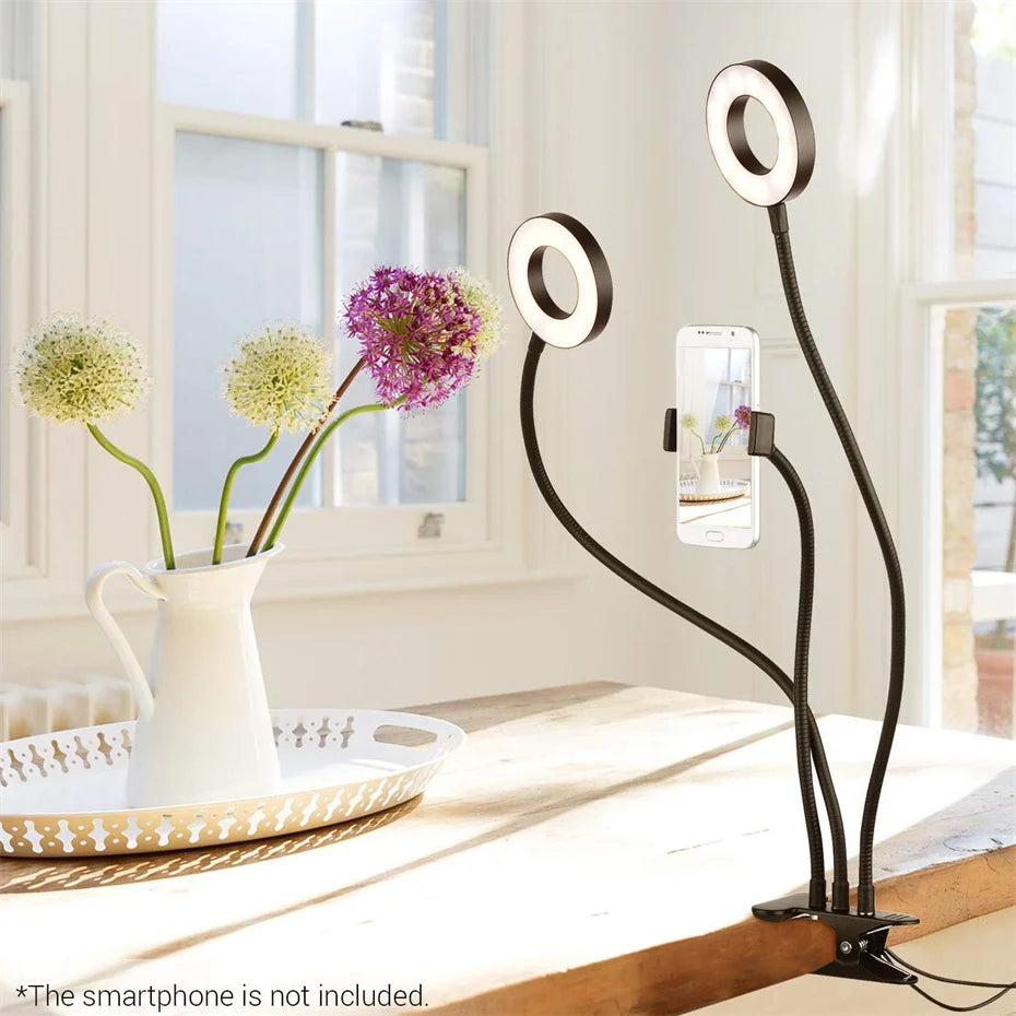 Black dual-arm desk lamp with adjustable circular LED ring lights and a central smartphone holder clamp.