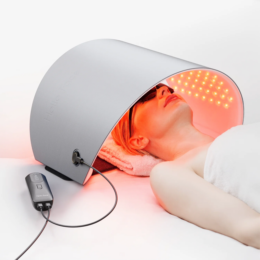 A person lying under a curved silver Hello Face LED light therapy mask emitting red and near-infrared light.