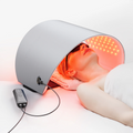 A person lying under a curved silver Hello Face LED light therapy mask emitting red and near-infrared light.