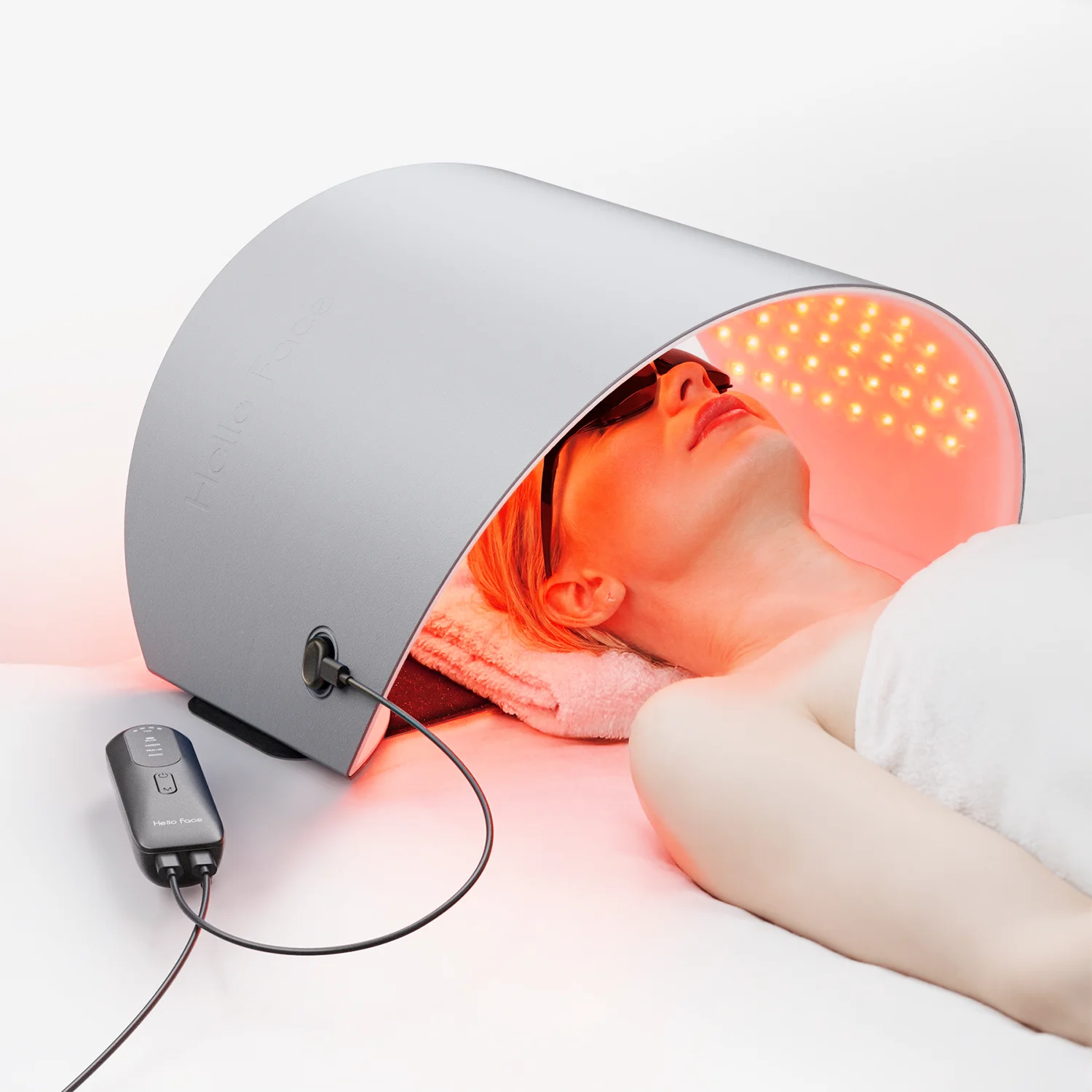 A person lying under a curved silver Hello Face LED light therapy mask emitting red and near-infrared light.