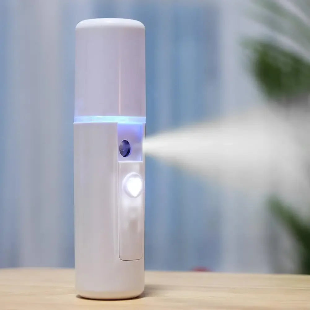 A portable white cylindrical facial mist sprayer standing upright on a wooden table, emitting a fine white mist.